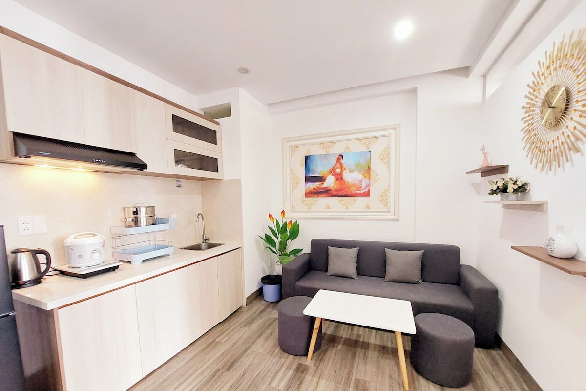 A modern kitchen with a built-in oven, a coffee maker, and a sink. There is a gray sofa with a white coffee table and a potted plant. A decorative wall piece is above the sofa.