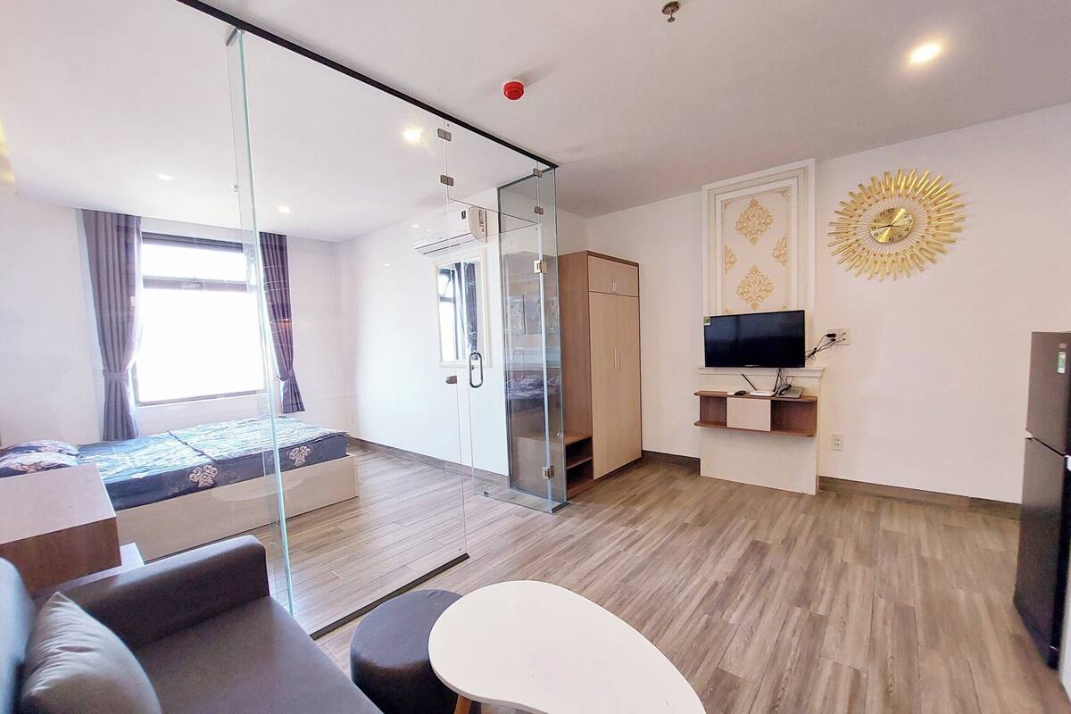 A modern hotel room with a glass partition, a bed, a sofa, a small table, a TV, and a wardrobe.