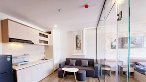 A modern kitchen with white cabinets, a built-in oven, and a refrigerator. There is a gray sofa with a round coffee table in the living area, and a glass partition separates the kitchen from the dining area. - Lavender II Hotel (Da Nang)