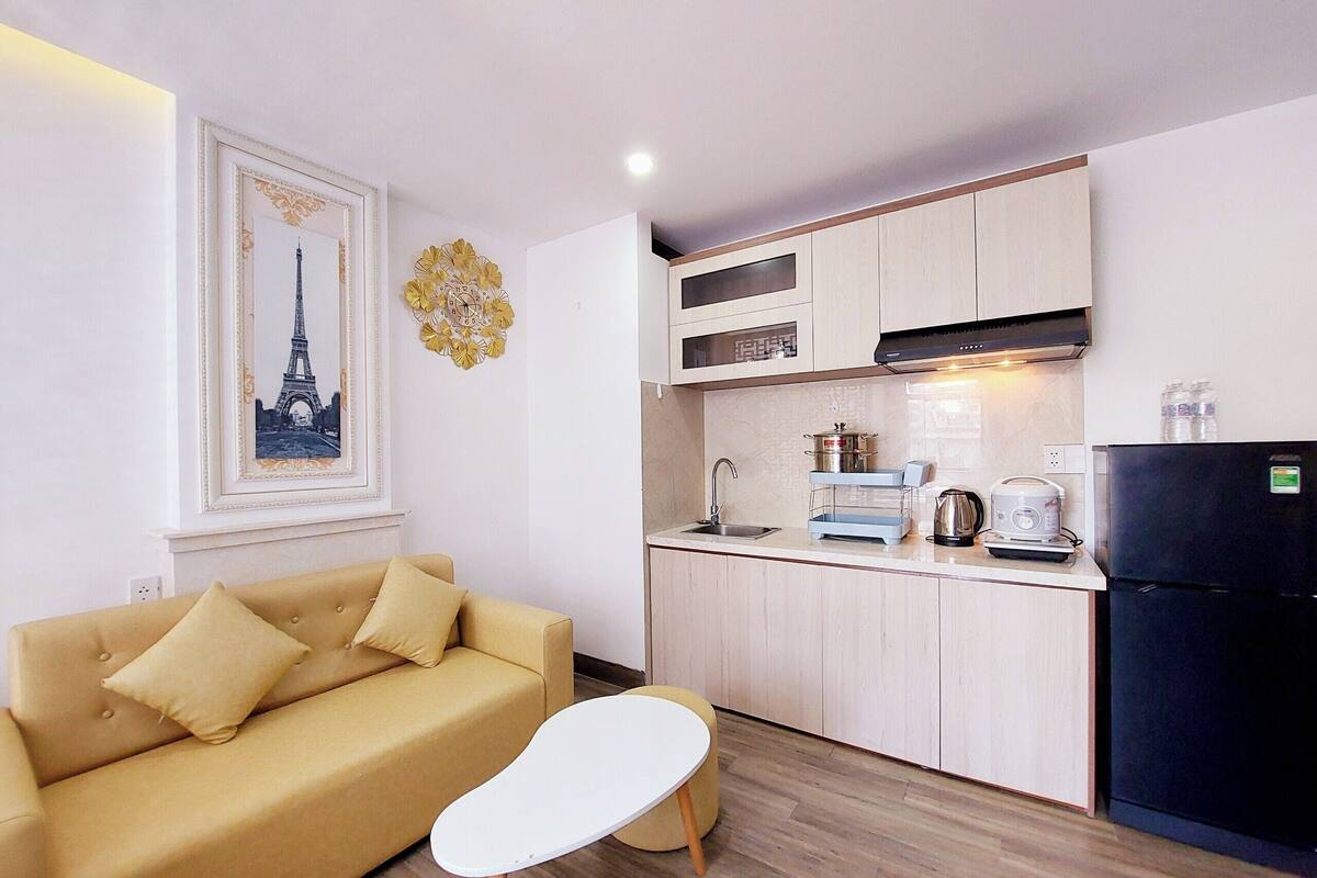 A modern kitchen with a built-in stove, a black refrigerator, and a sink. There is a beige sofa with two cushions and a small white table. A decorative clock and a framed picture of the Eiffel Tower are on the wall.