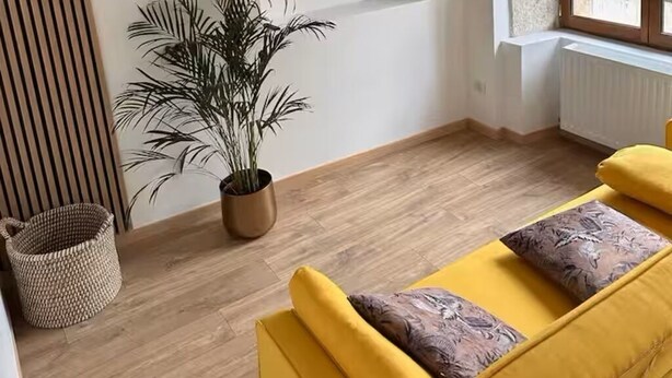 Ein modernes Wohnzimmer mit Holzboden, einem Wand-Fernseher, einer Zimmerpflanze, einem gelben Sofa mit gemusterten Kissen und einem Korbwäschesammler.