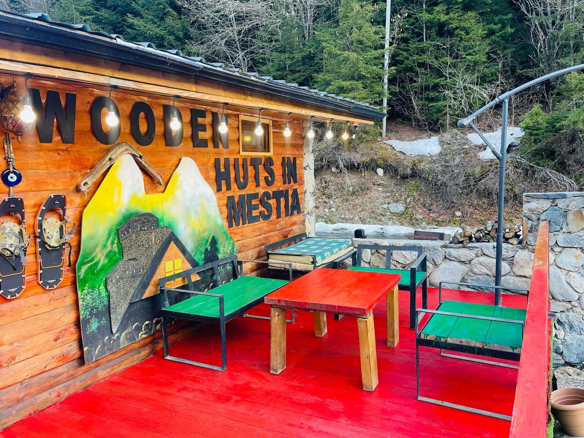Une cabane en bois sur laquelle est accrochée une pancarte indiquant « Cabanes en bois à Mesta ». Elle dispose d’un espace extérieur avec des tables et des bancs rouges et verts.