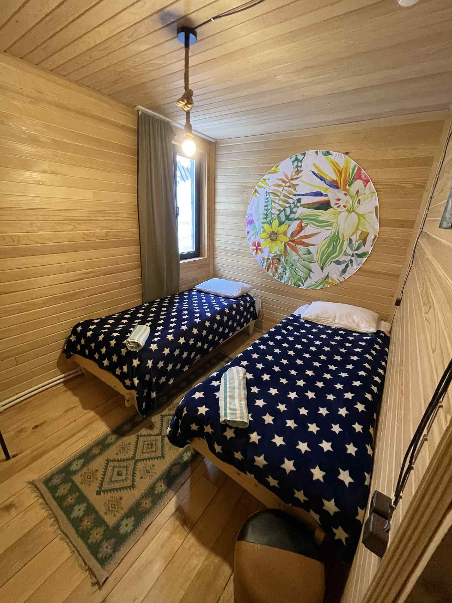 Une petite pièce agréable et chaleureuse, avec deux lits, une fenêtre avec des rideaux, un luminaire suspendu et un élément de décoration circulaire sur le mur.