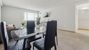 A modern, well-lit living room with a glass-top dining table, black chairs, a beige sofa, a potted plant, and a flat-screen TV mounted on the wall. - Legacy at Imperial Village 1108 I 1 Bed 1 Bath (Ric hmond)