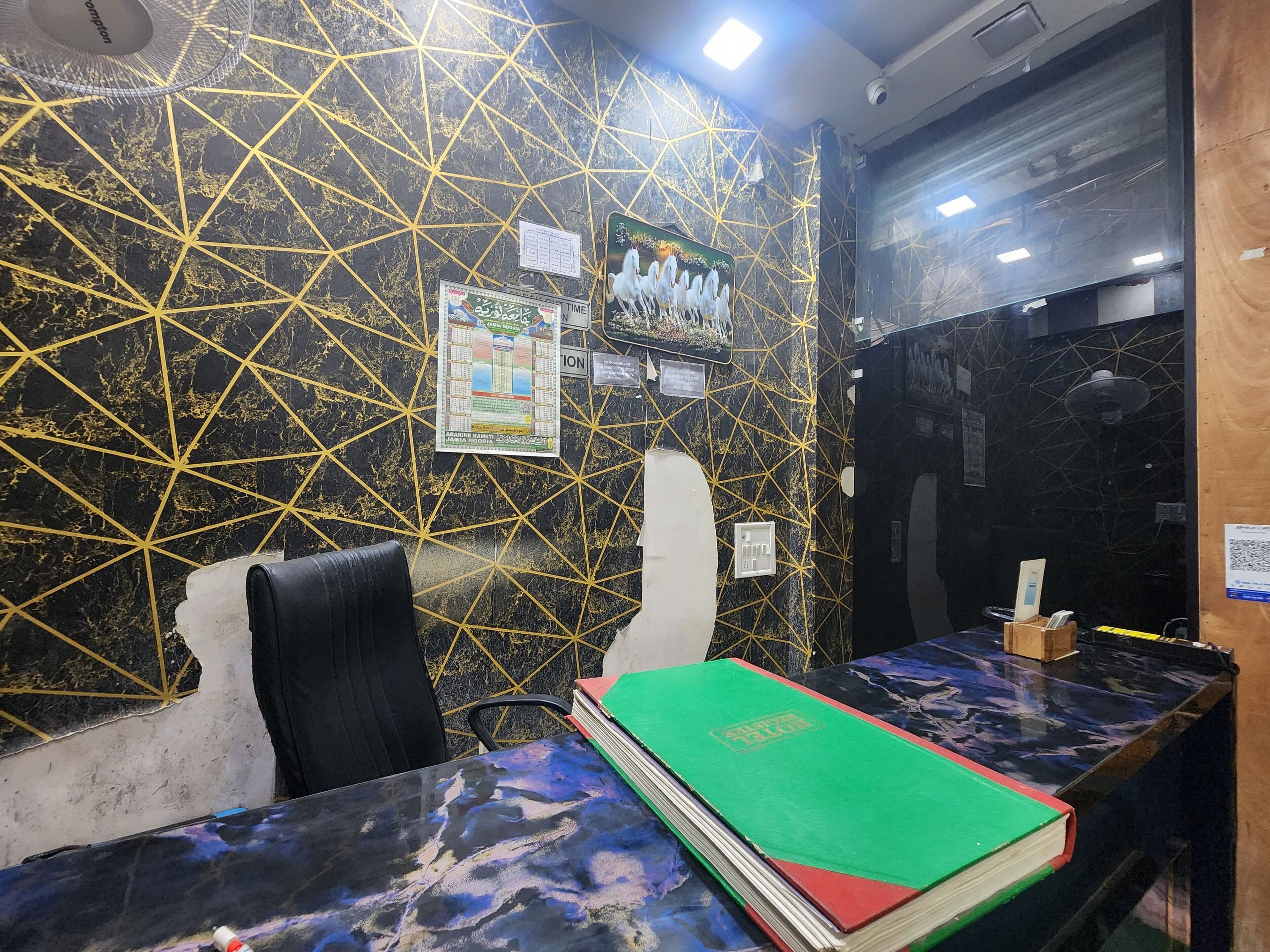 A hotel reception area with a marble wall featuring geometric patterns, a reception desk with a green book, and a black chair.