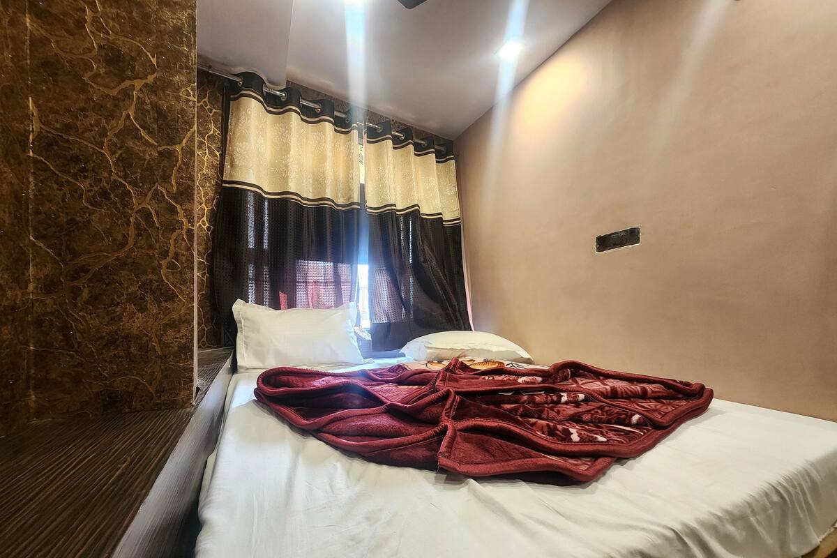 Hotellihuone, jossa on sänky, punainen peitto, valkoiset tyynyt ja kukkakuviollinen verho.