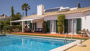 Een zwembad met een terras, een overdekt zitgedeelte en een gebouw met groene luiken en zonnepanelen.