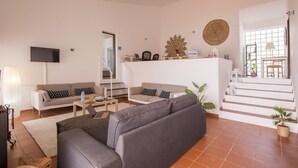 Een ruime woonkamer met een grijze bank, een salontafel en een aan de muur bevestigde televisie. Er is een trap die leidt naar een hoger gelegen verdieping met uitzicht op buiten.