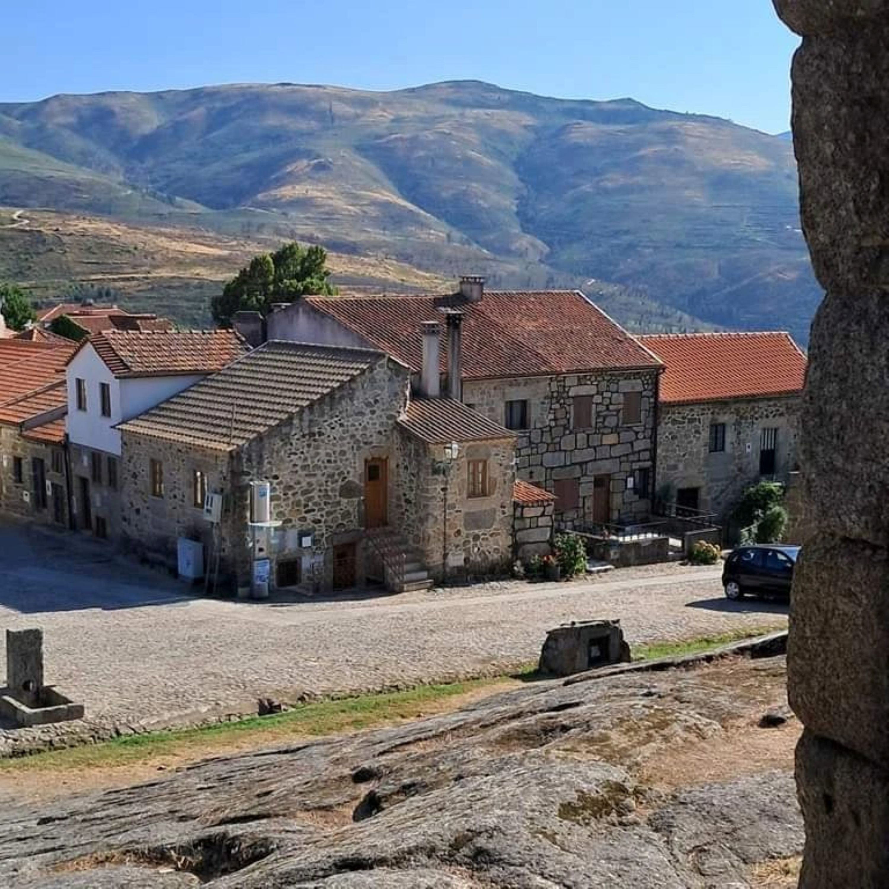 A stone building with a red-tiled roof, situated in a rural area with mountains in the background.