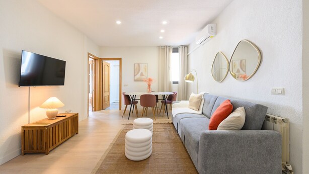 A modern living room with a flat-screen TV, a wooden console table with a lamp, a grey sofa with cushions, a dining area with a table and chairs, and two wall-mounted mirrors. - Two bedroom flat-Terrace and balcony in Madrid (Madrid)