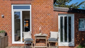 Ein Ziegelhaus mit einer Holzdachterrasse, zwei Klappstühlen, einem Tisch und einer Tür mit Glasfeldern.