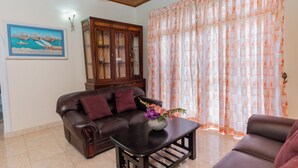 A living room with a brown leather sofa, a wooden cabinet, a coffee table with a vase of flowers, and a large window with orange curtains. - 4-bedroom Green Oasis in Kandy (Kandy)