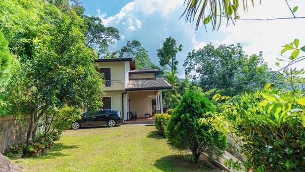 A two-story house with a covered porch, surrounded by lush greenery and a parked car. - 4-bedroom Green Oasis in Kandy (Kandy)