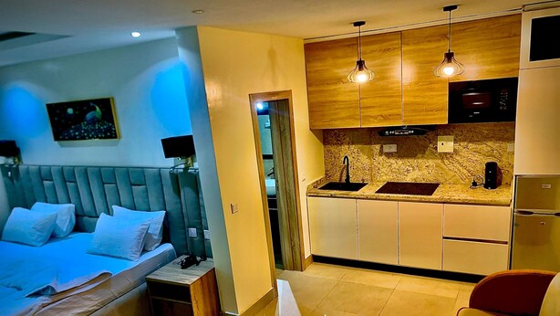 A compact hotel room with a bed, bedside table, and a small kitchenette. - Slavan Suites & Apartments (Owerri)