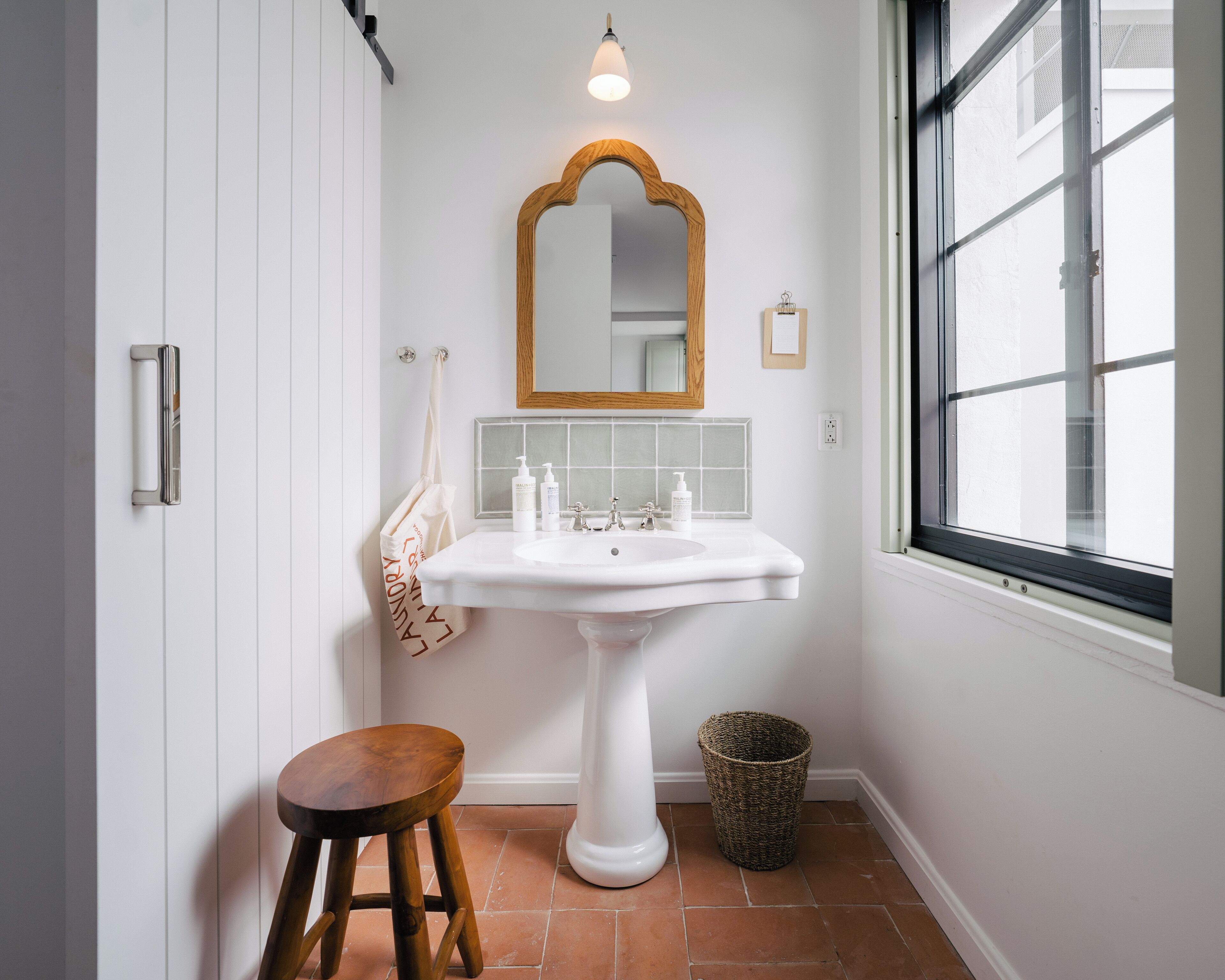 A bathroom with a white pedestal sink, a wooden stool, a mirror with a wooden frame, and a window.