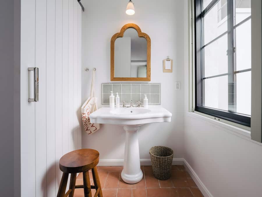 A bathroom with a white pedestal sink, a wooden stool, a mirror with a wooden frame, and a window.