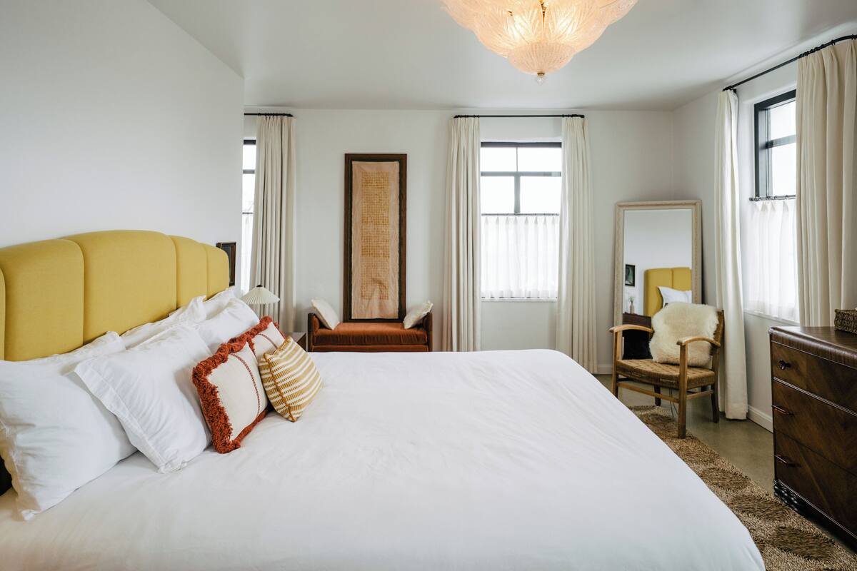 Una cama grande con ropa de cama blanca, un cabecero amarillo y varias almohadas. Hay una silla de madera y un aparador en la habitación.