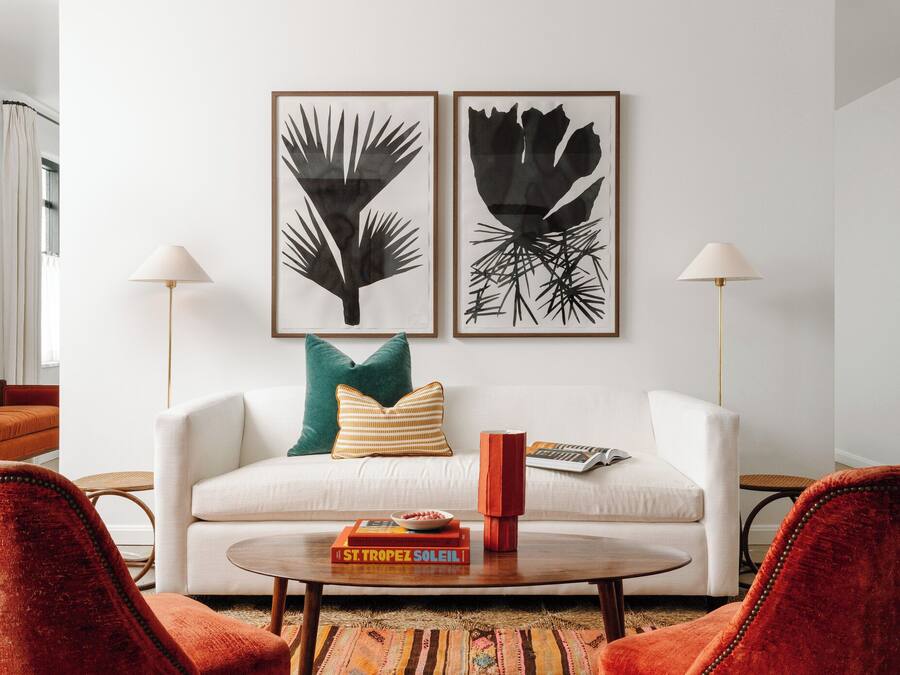 Una sala moderna con un sofá blanco, dos sillones rojos, un tapiz estampado y dos cuadros abstractos enmarcados en la pared.