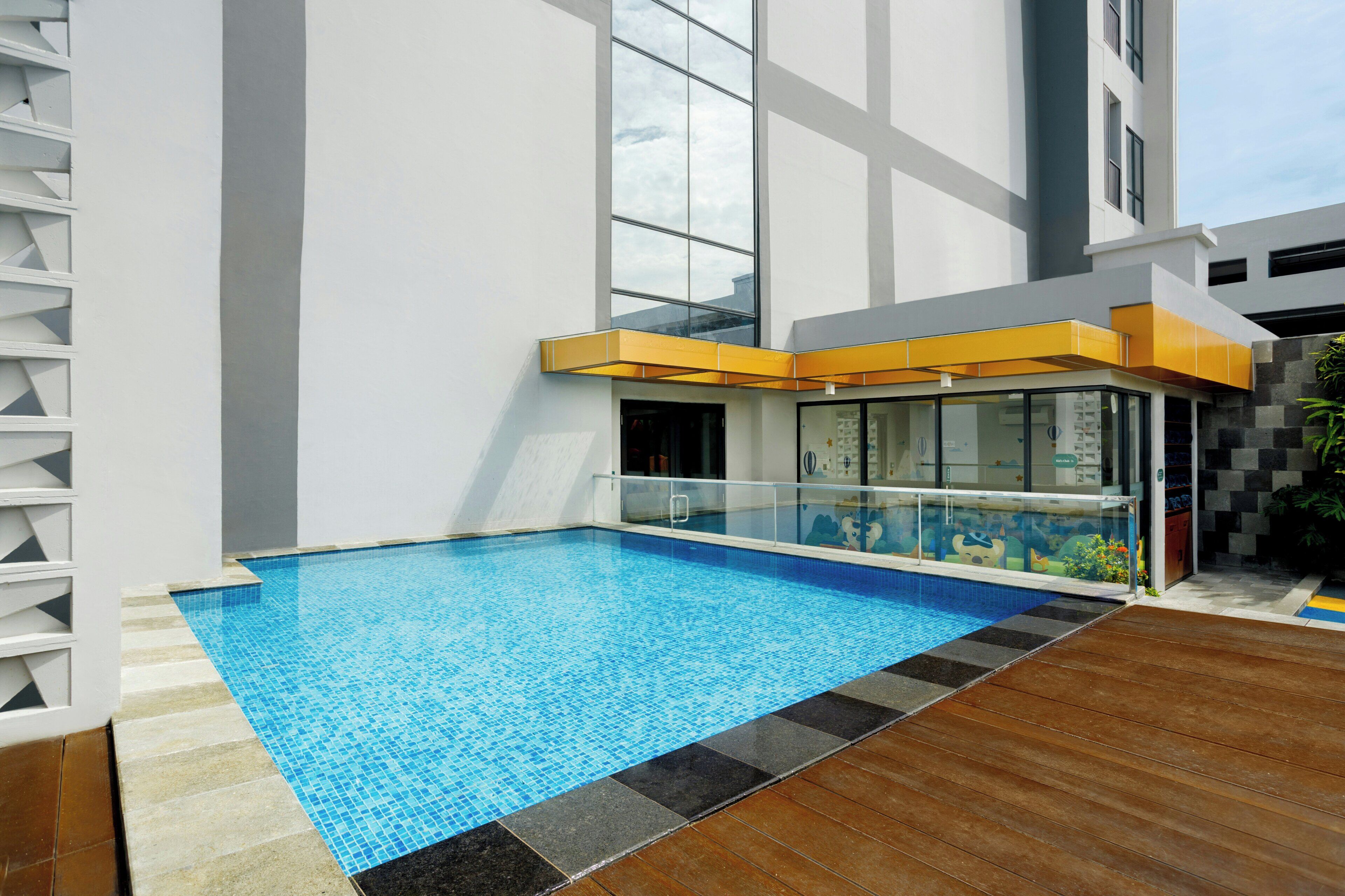 A modern indoor swimming pool with a clear blue water feature, surrounded by a wooden deck and a white building with large windows.