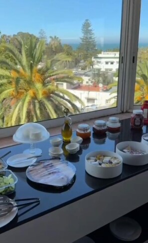 A dining table with various dishes and condiments, a window with a view of palm trees and buildings, and a clear sky.