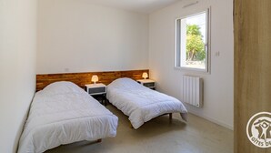 Una habitación pequeña y limpia con dos camas individuales, cabeceros de madera, mesitas de noche con lámparas, una ventana con vistas a zonas verdes y un radiador.