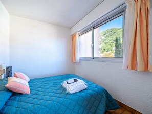 A bedroom with a blue quilted bedspread, a white mattress, and a view of a green landscape through a window with orange curtains.