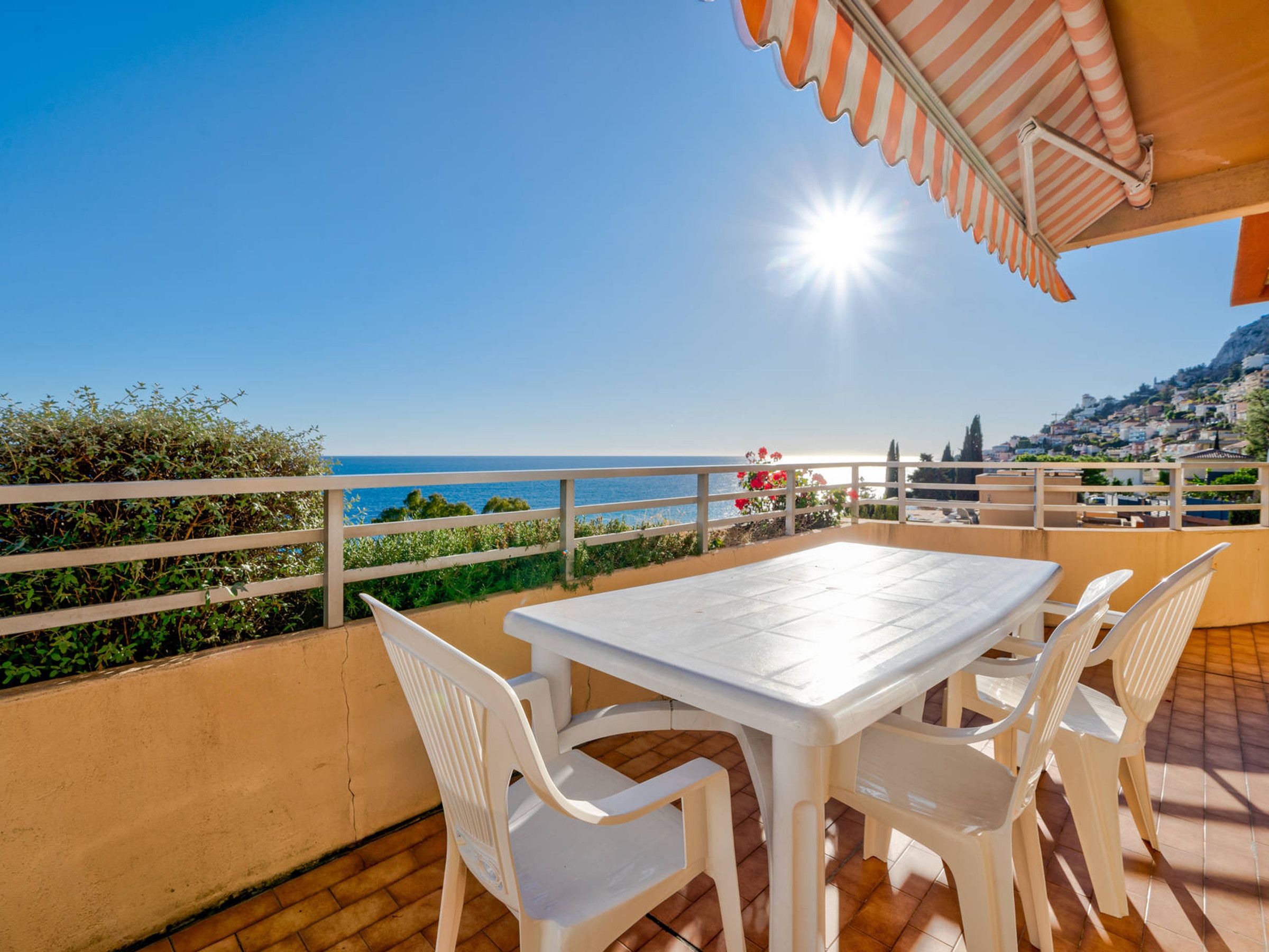 A terrace with a white table and chairs, overlooking a coastal view with a clear sky and sun.