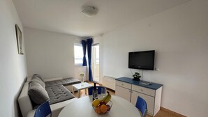 Een knus woongedeelte met een bank, een eettafel met stoelen, een kleine keukenhoek en een aan de muur bevestigde televisie.