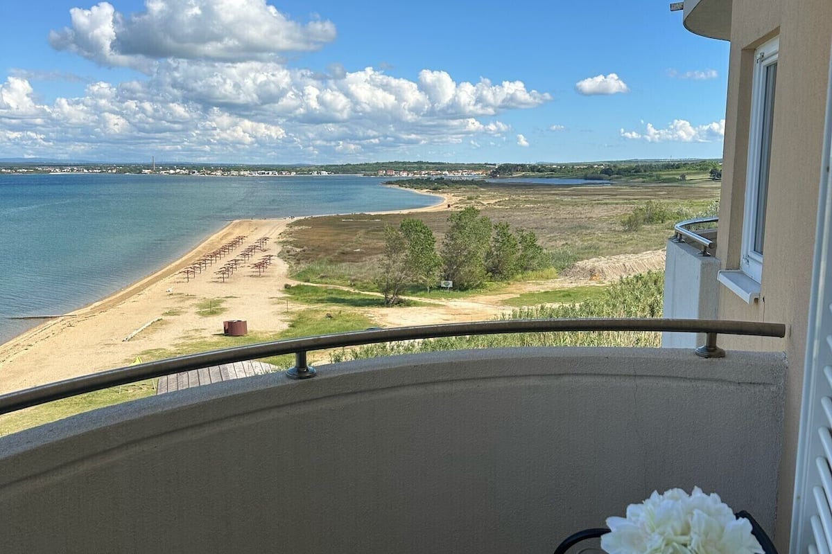A balcony with a view of a beach and water. There is a table with a bottle and a glass.