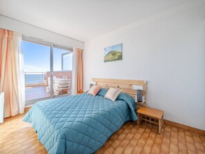 A bedroom with a large bed, a wooden headboard, a bedside table with a lamp, and a view of the sea through a sliding door.