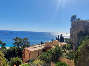 A coastal view with buildings overlooking the sea, a cliff with vegetation, and boats in the water.