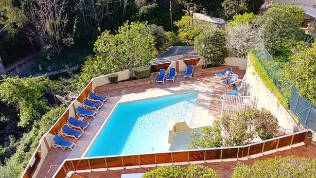 Une piscine entourée de chaises longues bleues, de verdure et d’une zone délimitée par une clôture.