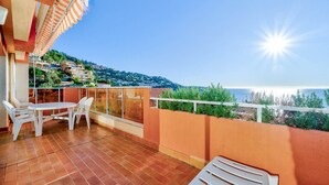 Une terrasse avec une table et des chaises, une balustrade, et une vue sur la mer et les bâtiments en arrière-plan.