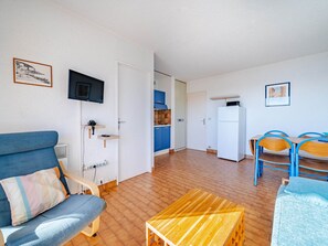A compact living space with a sofa, a small wooden table, a TV mounted on the wall, a kitchen area with blue cabinets, and a door leading to another room.