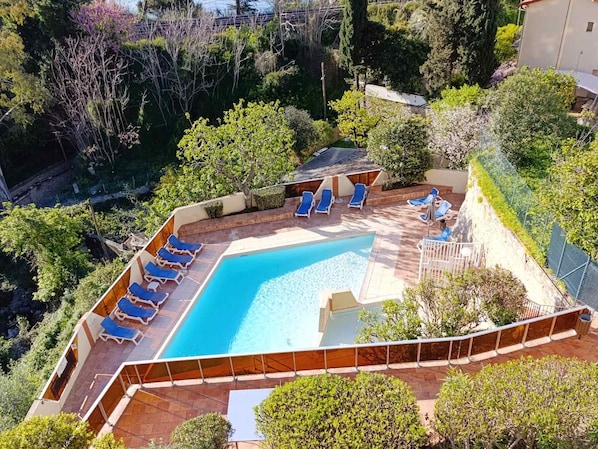 Piscina com espreguiçadeiras azuis, cercada por vegetação e uma área delimitada por cerca.