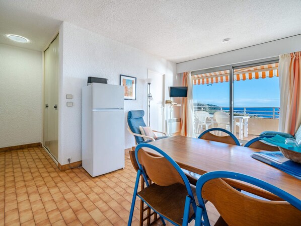 A compact room with a dining table, chairs, a refrigerator, and a sliding glass door leading to an outdoor area with a view of the sea.