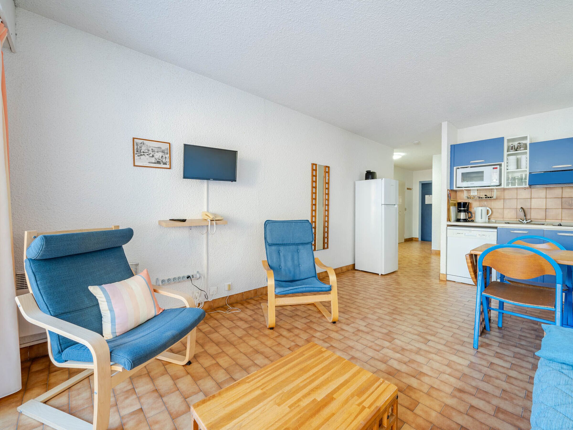 Un salon avec deux fauteuils bleus, une table basse en bois, un téléviseur à écran plat et un coin cuisine doté de meubles bleus et d’un four à micro-ondes.