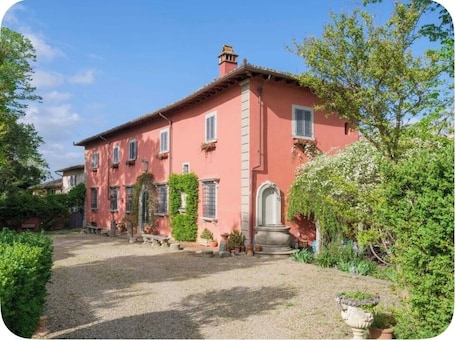 A pink villa with a gravel driveway, surrounded by greenery and potted plants.