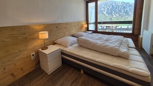 Un dormitorio con cabecera de madera, una cama grande con colchón, dos lámparas de noche y una ventana con vistas a la montaña.