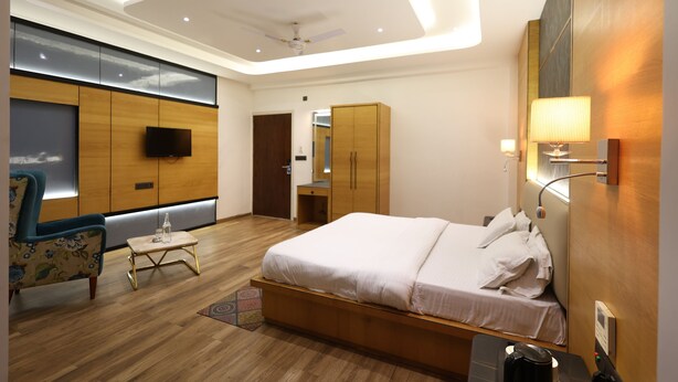 A modern hotel room with a large bed, a seating area with a chair and table, a wardrobe, and a television. - Vasuh Mhangglam (Bilha)
