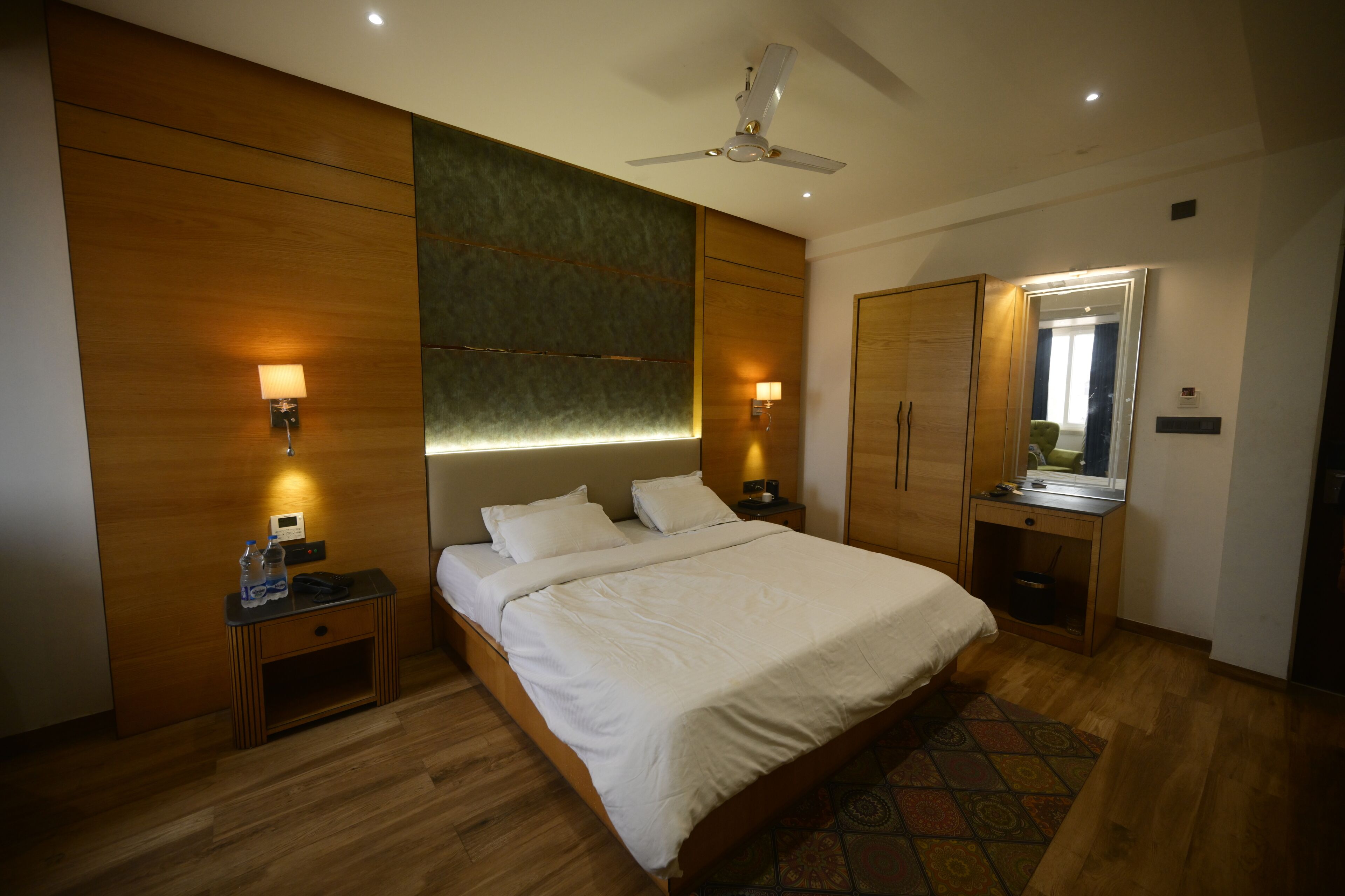 A modern hotel room with a large bed, wooden panel walls, a ceiling fan, bedside lamps, a small desk with a mirror, and a patterned rug.
