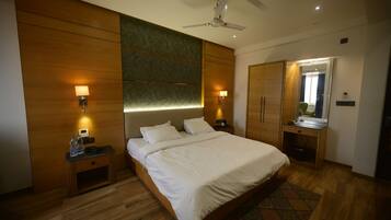 A modern hotel room with a large bed, wooden panel walls, a ceiling fan, bedside lamps, a small desk with a mirror, and a patterned rug.