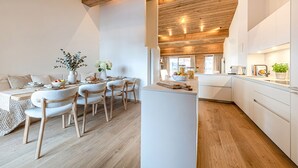 Et moderne køkken med trædetaljer, et spiseområde med hvide møbler og en hyggelig opholdsstue med sofa og dekorative planter.