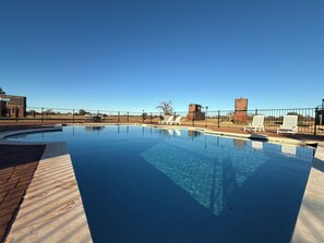 A rectangular swimming pool with a brick border, surrounded by a black fence. There are lounge chairs and a small building with a sign in the background. - Wine Country · Hot Tub, BBQ & Pool Near Wineries (Fredericksburg)