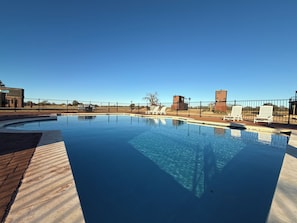 A rectangular swimming pool with a brick border, surrounded by a black fence. There are lounge chairs and a small building with a sign in the background.