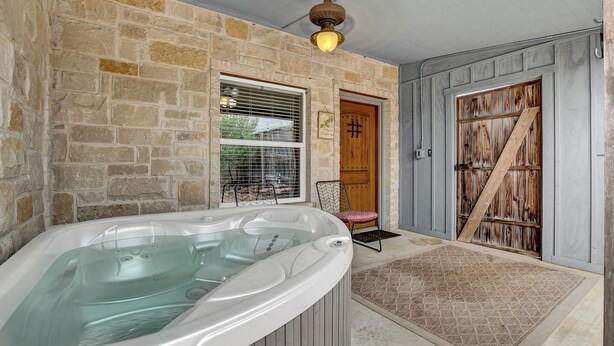 A stone-walled bathroom with a hot tub, a wooden door, and a window with blinds. - Wine Train Cottage · Hot Tub, Pool & Wine Trail (Fredericksburg)