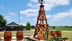 A rustic windmill structure surrounded by wooden barrels and desert plants, with a barn and open fields in the background. - Wine Train Cottage · Hot Tub, Pool & Wine Trail (Fredericksburg)