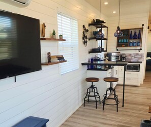 A compact kitchen with a flat-screen TV, wooden shelves, a coffee machine, and a small dining area with stools.