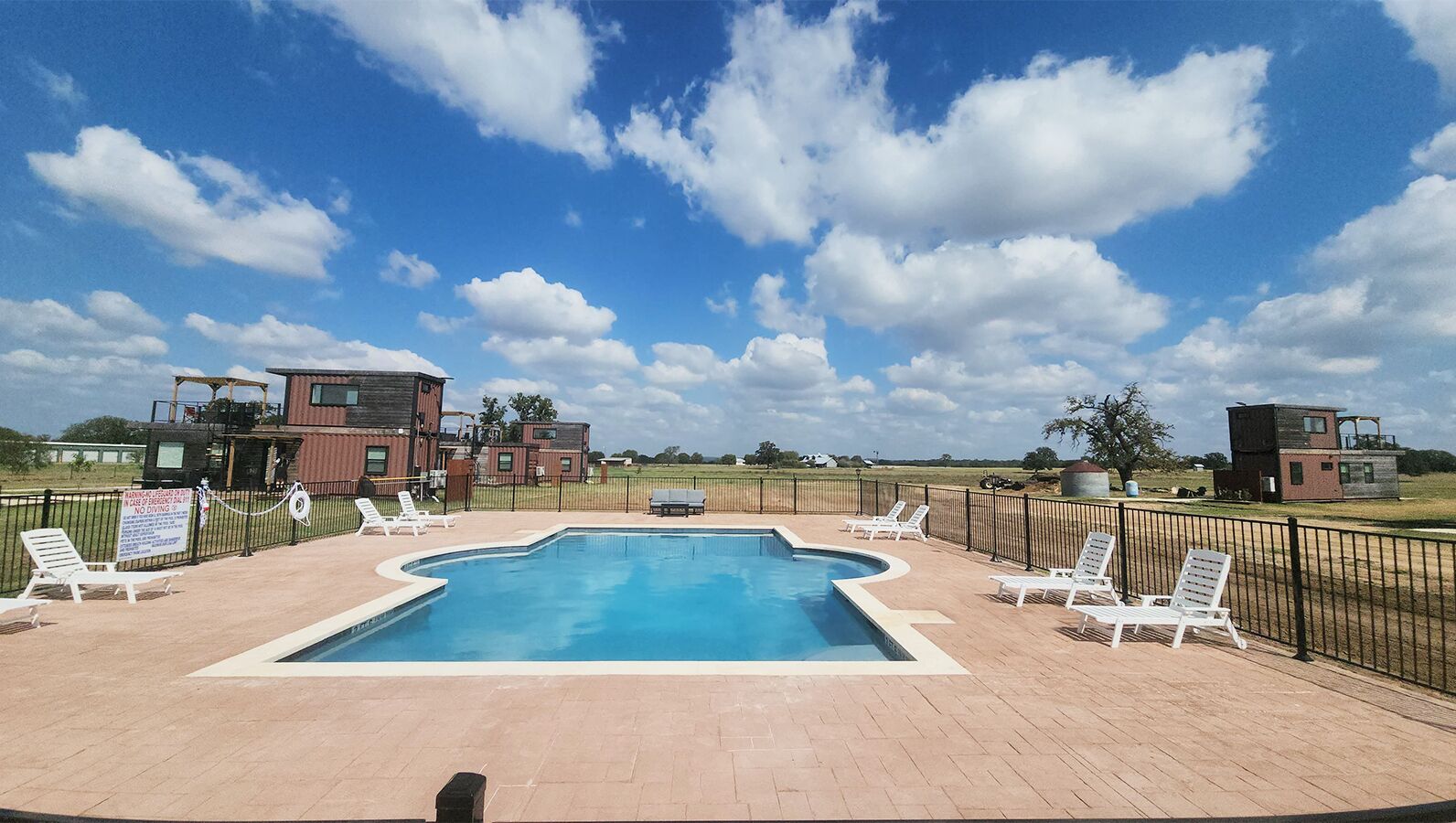 Une piscine rectangulaire, bordée d’une terrasse carrelée, entourée de chaises longues blanches et d’une clôture en métal noir.