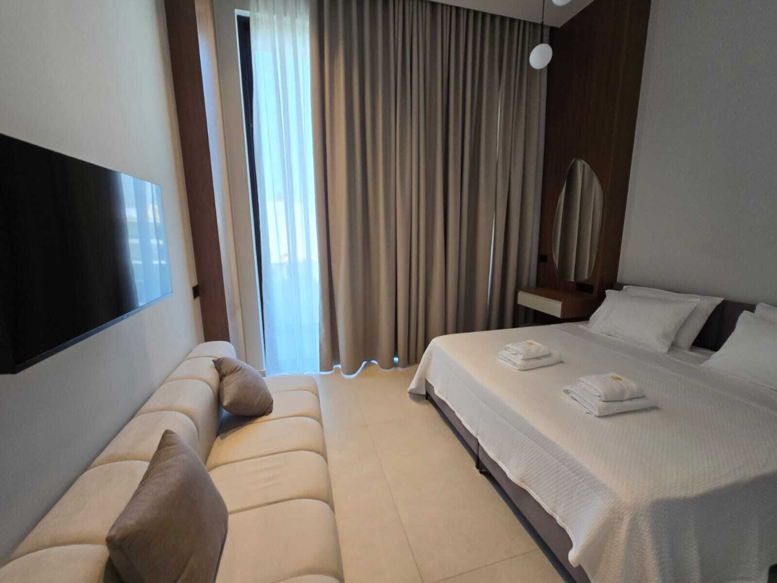 A modern hotel room with a large bed, a sofa, a flat-screen TV, and a view of the outdoors through a window with curtains.